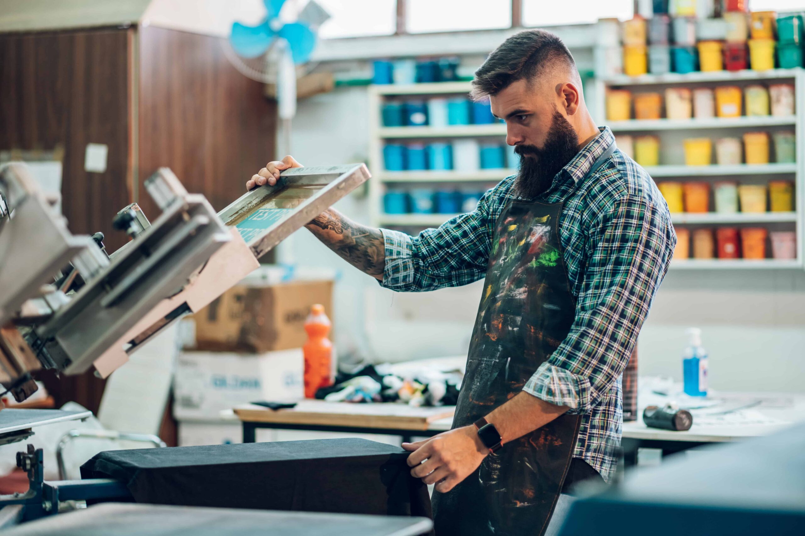 A bearded man wearing a paint-splattered apron performs Screen Printing Little Rock services by lifting a silkscreen frame from a black t-shirt in a professional print shop.