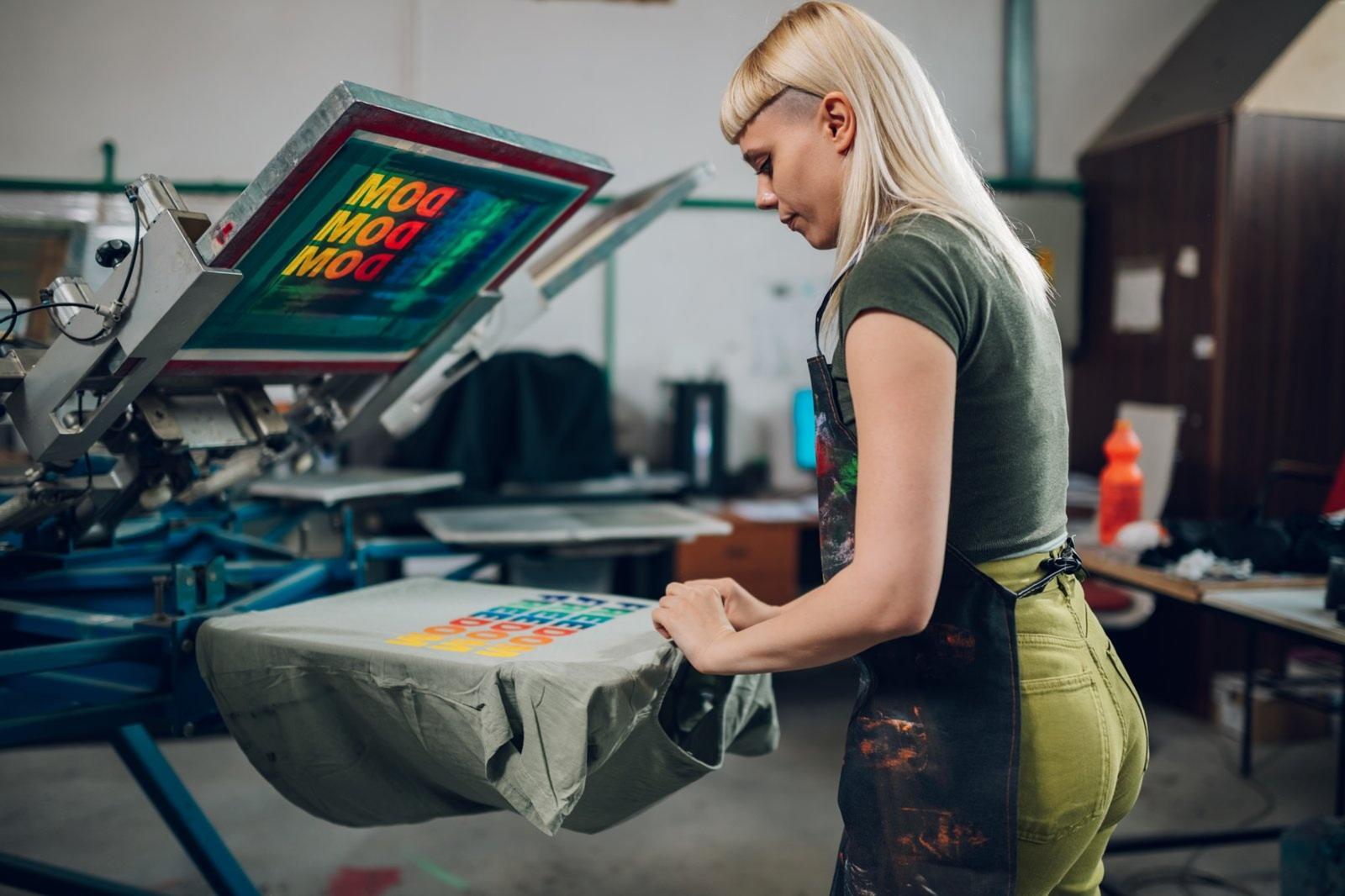 A female printmaker in a paint-splattered apron operates a professional screen press with a colorful "MOOD" graphic, showcasing the hands-on quality of Women's Affordable T-Shirt Printing for those seeking reliable women's affordable t shirt printing services and stylish women's custom tee shirts.