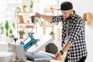 A male artisan in a plaid shirt operates a professional heat press machine in a bright, plant-filled printing shop, carefully aligning a white garment to deliver top-quality Affordable T-Shirt Printing Services for clients seeking reliable t shirt printing and custom made t shirt shirt printing.