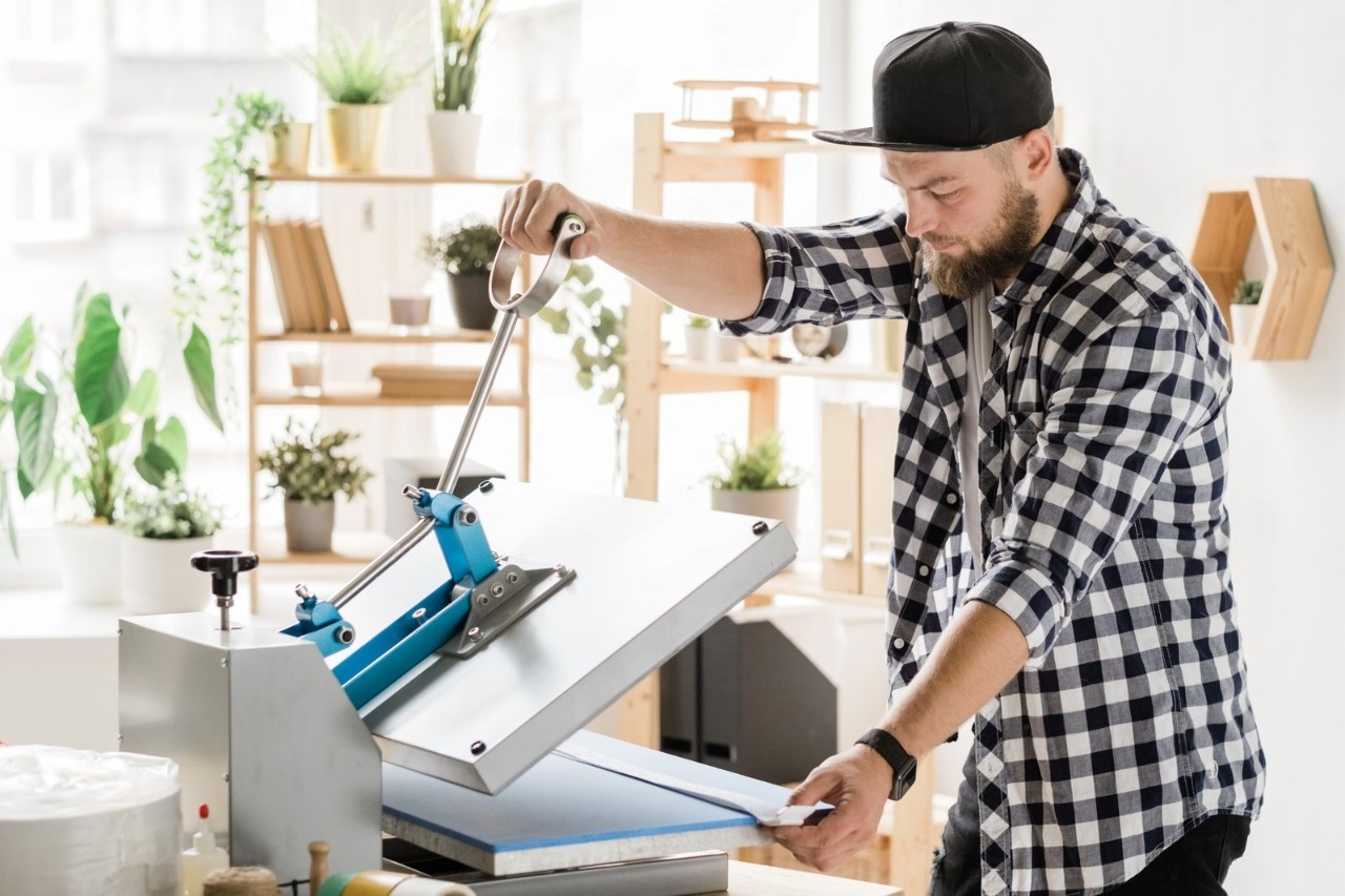 A male artisan in a plaid shirt operates a professional heat press machine in a bright, plant-filled printing shop, carefully aligning a white garment to deliver top-quality Affordable T-Shirt Printing Services for clients seeking reliable t shirt printing and custom made t shirt shirt printing.