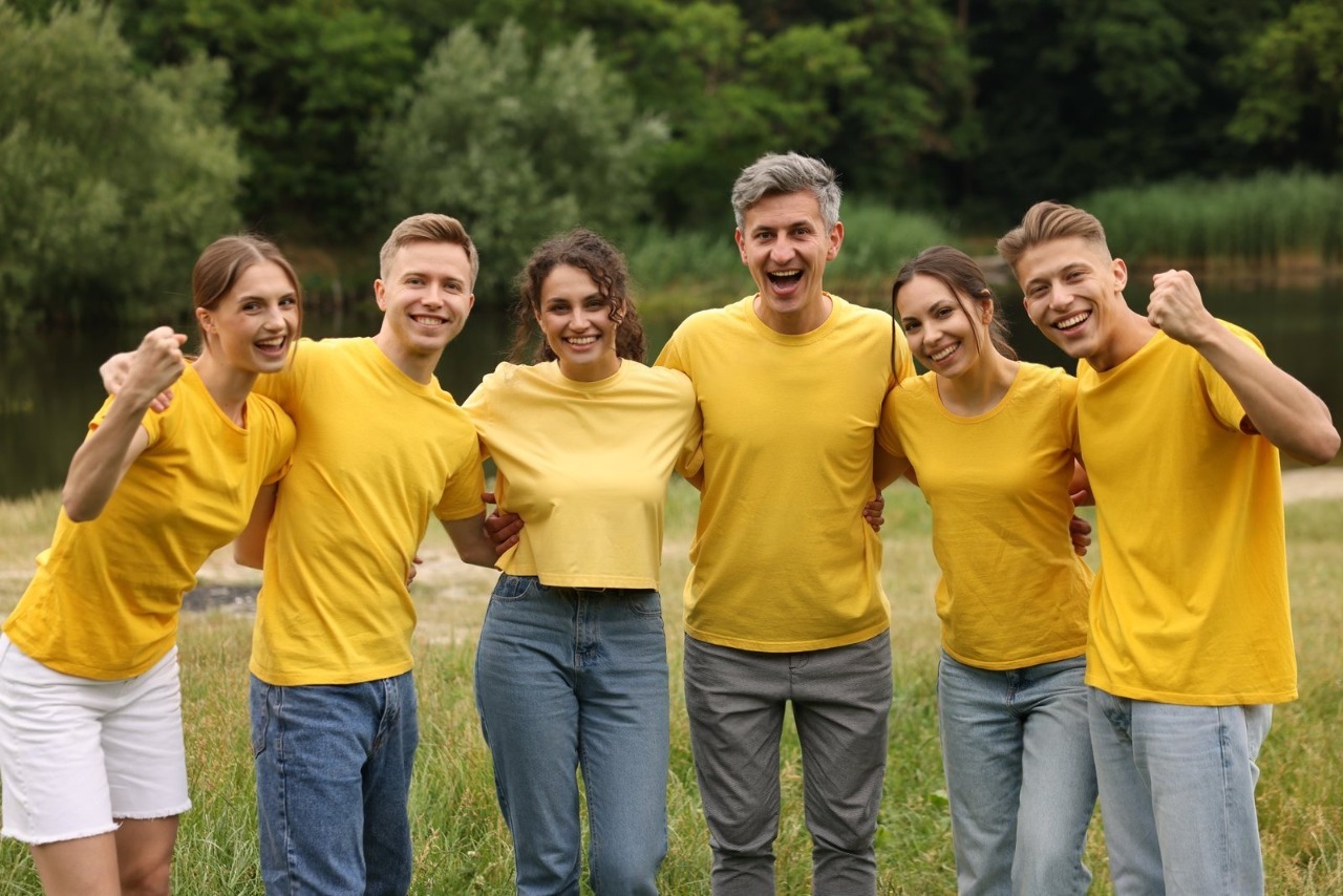 A joyful group of six family members standing arm-in-arm outdoors by a lake, all wearing vibrant matching yellow t-shirts and cheering, perfectly showcasing the fun and unity created by custom family reunion shirts little rock.