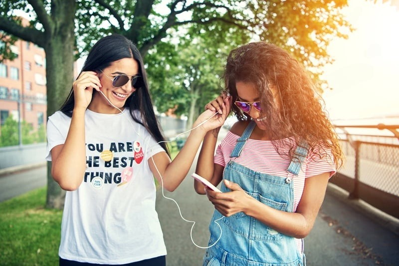 Two smiling young women sharing earphones outdoors, with one wearing a custom graphic tee reading "YOU ARE THE BEST THING," showcasing the vibrant, personalized quality of Girl's Affordable T-Shirt Printing available through a dedicated local girl's affordable t-shirt printing service.