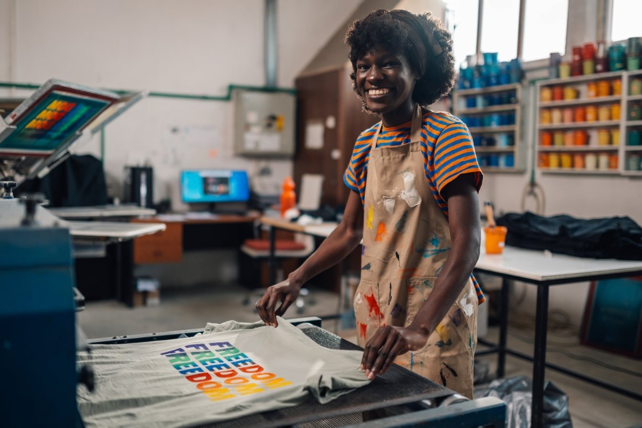 A smiling female artisan in a paint-splattered apron operates a professional screen printing press in a bright studio, showcasing the high-quality, vibrant results you get with dedicated Kid's Affordable T-Shirt Printing for your kids custom shirts and youth t shirt printing needs.