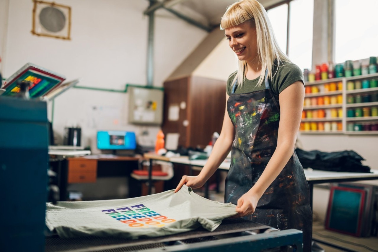 A smiling female printmaker wearing a paint-splattered apron admires a freshly printed custom graphic tee in a vibrant workshop, showcasing the expert craftsmanship and local dedication of a premier Little Rock T-Shirt Company ready to fulfill your bulk t shirt printing little rock orders.