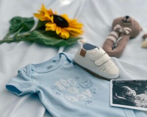 A flat lay photo on a soft white sheet showing a light blue baby onesie embroidered with "It's a boy", a small white infant sneaker, an ultrasound scan photo, a knitted teddy rattle, and a bright sunflower, representing the inspiration behind creating custom baby shower shirts for guests.