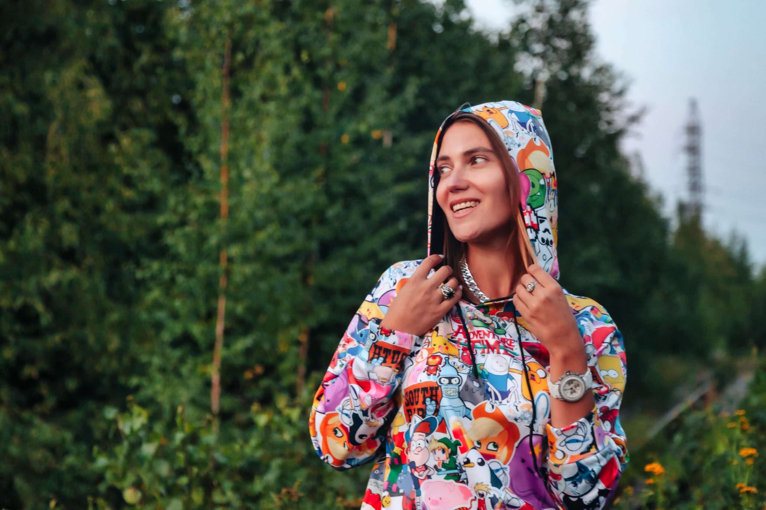 A young woman smiling outdoors while wearing a vibrant, multicolored cartoon character collage hoodie with the hood pulled up, showcasing premium, edge-to-edge ink coverage options for custom hoodies little rock arkansas.