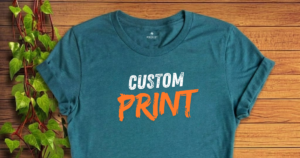 A soft, dark teal t-shirt featuring the text "CUSTOM PRINT" in bold white and orange lettering, laid flat on a wooden surface next to a green ivy vine, highlighting the premium quality of our graduation shirts little rock.