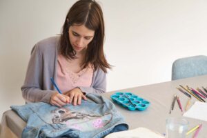A focused artist sitting at a table and hand-painting a detailed deer design onto a denim jacket, illustrating the meticulous craftsmanship behind custom apparel, providing a prime context for discussing airbrushing on a shirt and airbrush shirt durability.