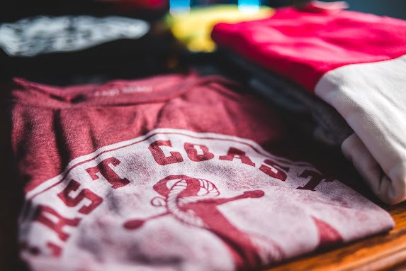 A close-up, shallow depth-of-field view of folded custom t-shirts stacked on a wooden surface, with the front maroon shirt showing a white circular "EAST COAST" anchor graphic, highlighting the clarity of professional garment decoration and factors impacting the final T-Shirt Printing Cost.