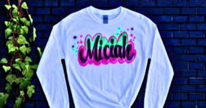 A white long-sleeve shirt displaying the name "Miciah" in bold black cursive with vibrant neon pink and green airbrushed highlights, demonstrating the stunning artistry of custom memorial airbrush shirts little rock and airbrush tribute shirts little rock.