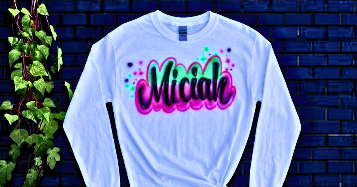 A white long-sleeve shirt displaying the name "Miciah" in bold black cursive with vibrant neon pink and green airbrushed highlights, demonstrating the stunning artistry of custom memorial airbrush shirts little rock and airbrush tribute shirts little rock.