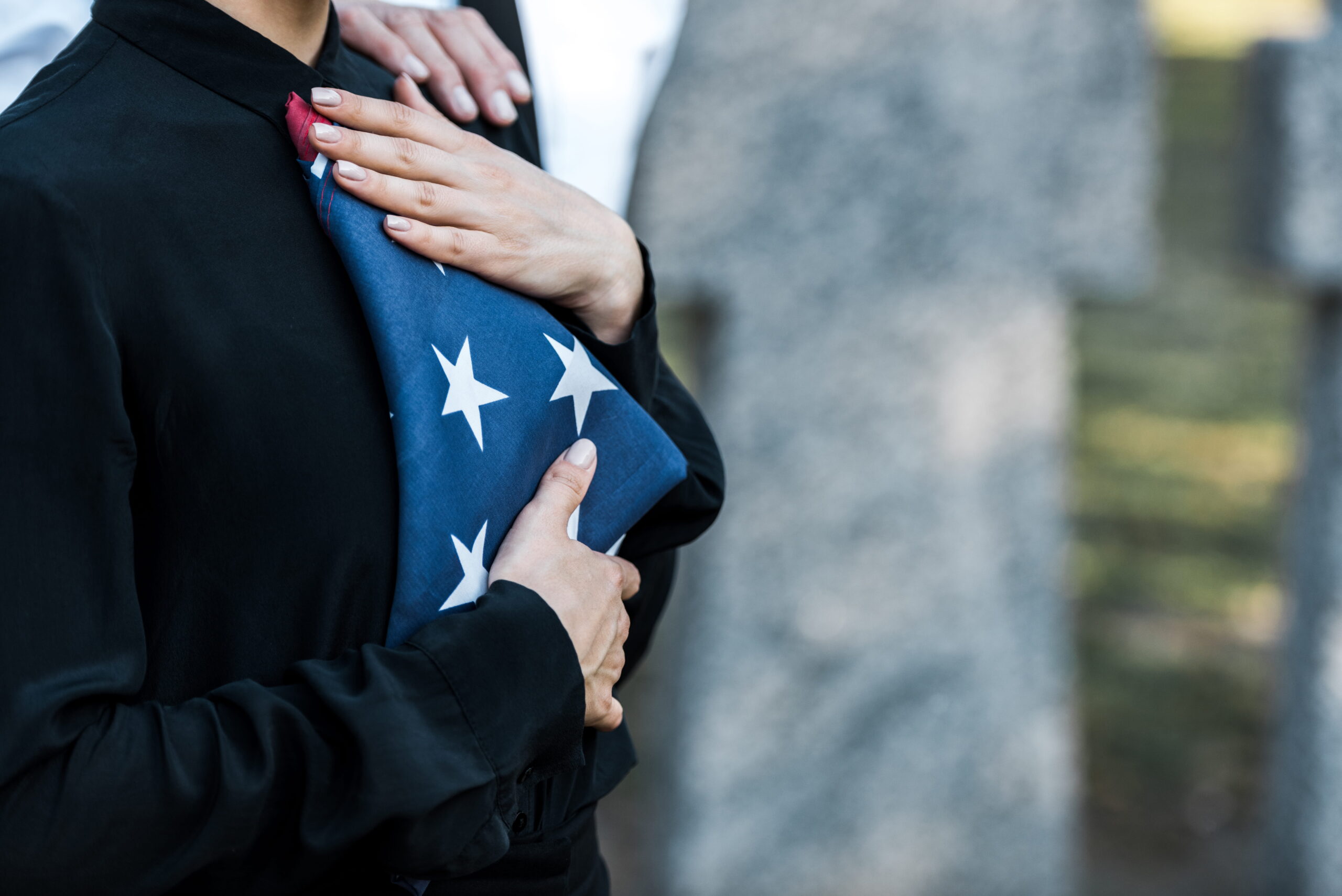 A close-up side profile of a mourning individual wearing a black button-down shirt, reverently holding a folded American burial flag to their chest at a graveside service, prompting families to ask What Is RIP Shirt customization options and how they can create a personalized wearable tribute.
