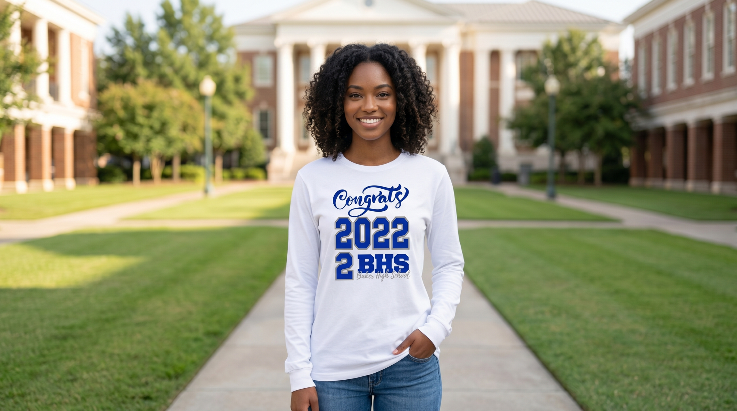 A smiling young Black woman with beautiful curly hair stands on a brick campus pathway wearing a custom white long-sleeve t-shirt with a blue "Congrats 2022 BHS" design, showcasing premium options for women graduation shirts near me.