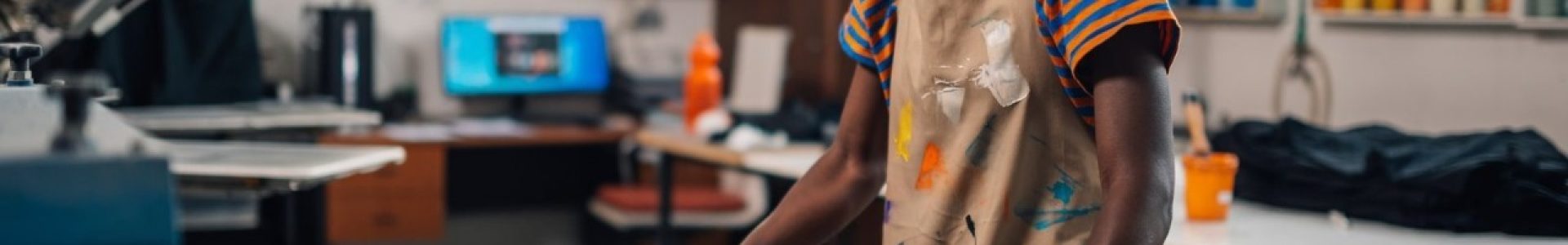 A smiling female artisan in a paint-splattered apron operates a professional screen printing press in a bright studio, showcasing the high-quality, vibrant results you get with dedicated Kid's Affordable T-Shirt Printing for your kids custom shirts and youth t shirt printing needs.