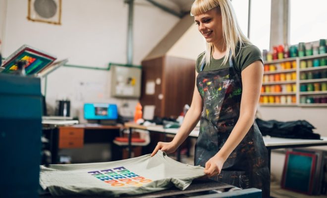 A smiling female printmaker wearing a paint-splattered apron admires a freshly printed custom graphic tee in a vibrant workshop, showcasing the expert craftsmanship and local dedication of a premier Little Rock T-Shirt Company ready to fulfill your bulk t shirt printing little rock orders.